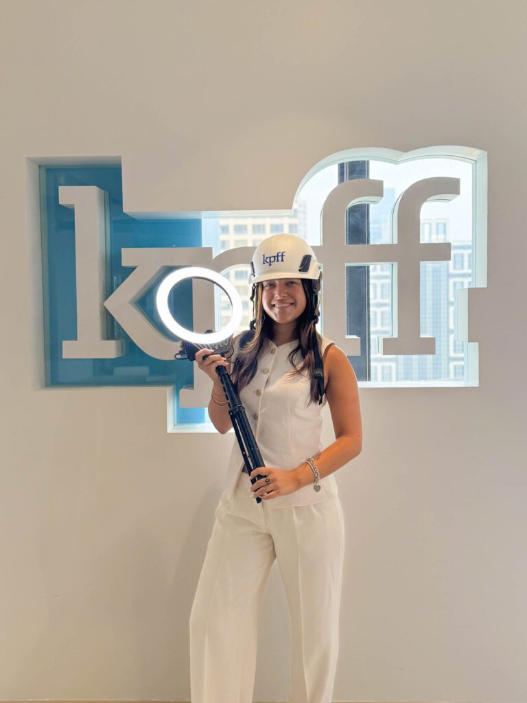 Social media intern stands in front of logo wall while wearing a logo helmet and holding a ring light. 