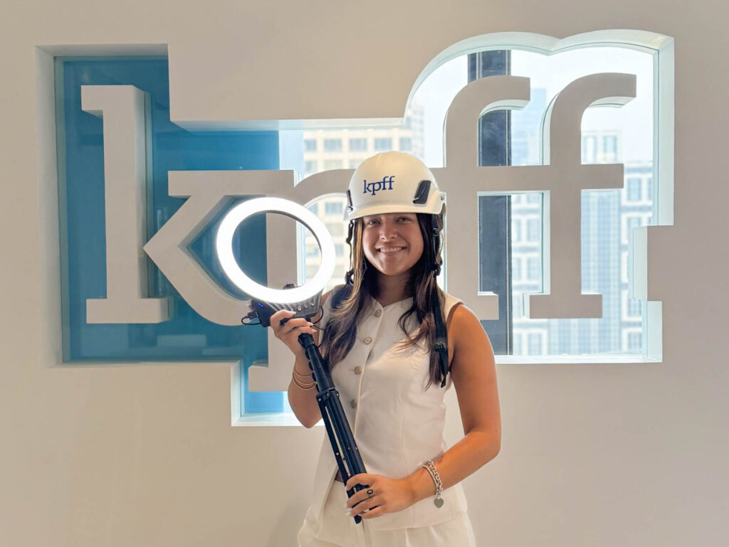 Social media intern stands in front of logo wall while wearing a logo helmet and holding a ring light.